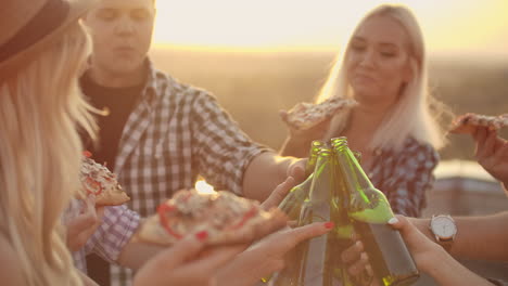 The-company-of-young-people-celebrating-a-friends-birthday.-They-eat-hot-pizza-and-drink-beer-and-enjoy-smiles-on-a-summer-evening-on-the-roof.-They-clink-beer-in-the-green-bottles.